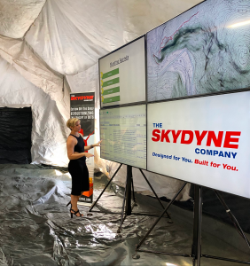 Inside an HDT Airbeam Tent, there is a woman standing in front of four 75" TVs mounted onto two Komodo Monitor Stands which make up a video or media wall.